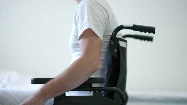 White bed in hospital ward with unrecognizable Caucasian man rolling wheelchair entering. Side view ill male patient with paralysis riding mobility aid device to window looking out. Illness and hope