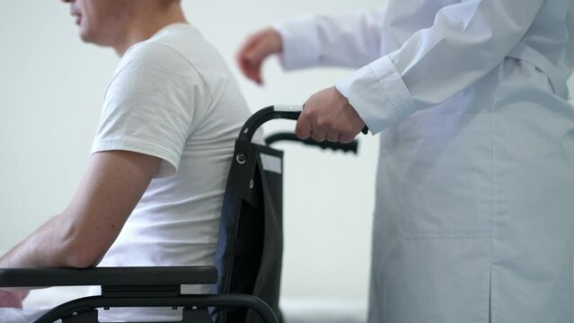 Side view nurse rolling wheelchair with patient entering white clean hospital ward. Unrecognizable ill Caucasian man in medical clinic supported by professional doctor helping. Accident rehabilitation