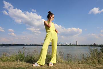 Happy slim girl in sunglasses and yellow summer suit standing on a beach on background of city buildings. Females fashion and beauty, travel and holidays in hot sunny weather