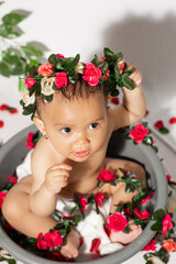 zenith shot of a beautiful latin baby girl with a flower crown, sitting on a gray cube and surrounded by red flowers. little brown-skinned girl looking up while holding her head.
