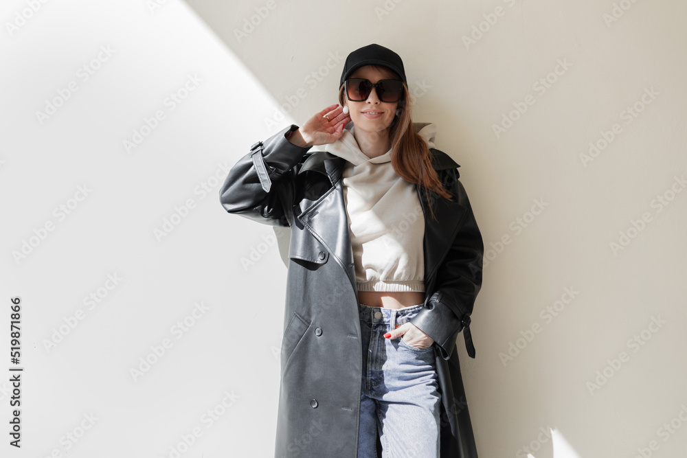 Sticker Beautiful fashion woman model with cap and trendy sunglasses in fashionable leather coat and hoodie with jeans stands near a wall with sunlight. Female urban clothes look