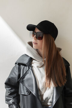 Cool fashionable hipster woman model with trendy sunglasses with black cap in leather black coat and hoodie stands near a wall in sunlight