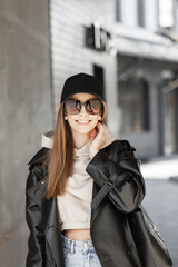 Fashionable happiness trendy woman with smile with black cap and trendy sunglasses in urban leather coat, jeans and hoodie with black leather bag walks on the street