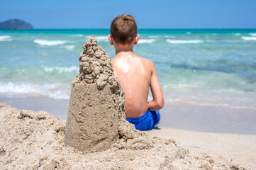 Kind sitzt an einem tropischen Sandstrand hinter einer Sandburg und schaut auf das türkisblaue Meer