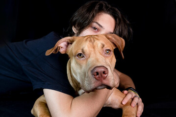 Portrait of a young man with a caramel colored pitbull dog against black background.