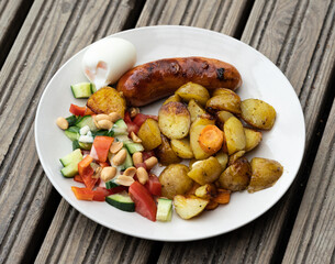 High angle view portion on a plate of delicious barbeque food on a wooden platform. Grilled sausage with rustic potato cubes, boiled egg and fresh salad.