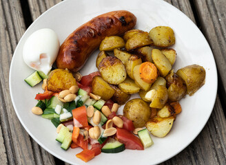High angle view portion on a plate of delicious barbeque food on a wooden platform. Grilled sausage with rustic potato cubes, boiled egg and fresh salad.