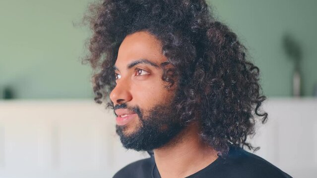 Close up young cheerful millennial middle eastern man with black curly hair smiling turns head and looks at screen located indoors at home or office. Positive emotions, sincerity, goodwill concept