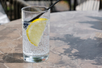 A glass of cold sparkling soda with a slice of yellow lemon on a table in the sun. Image with selective focus.