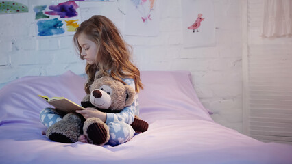 Side view of kid in pajama reading book and holding soft toy on bed at home.