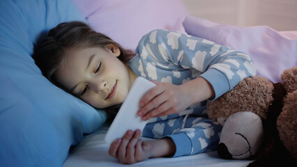 Preteen girl in pajama looking at smartphone near teddy bear on bed in evening.