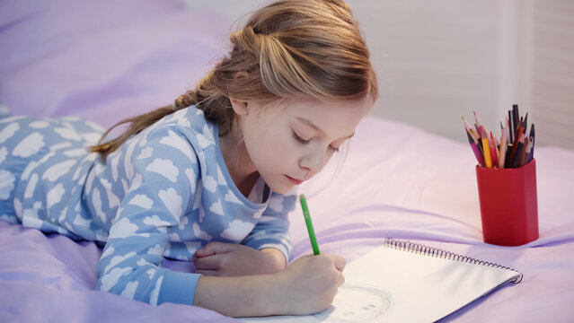 Preteen kid in pajama drawing on sketchbook on bed.