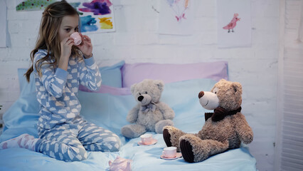 Preteen child drinking tea near soft toys on bed in evening.