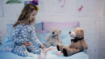 Preteen kid in pajama playing with tea cups near toys on bed.