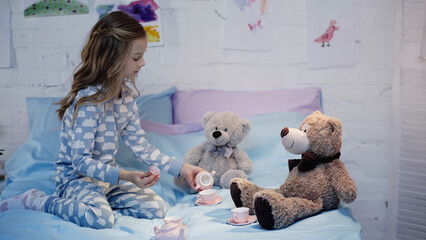Side view of preteen child in pajama playing near tea cups and soft toys on bed.