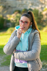 Fototapeta premium Retrato de chica joven con chaqueta gris y jersey en bosque al aire libre con pelo recogido y dedo en los labios