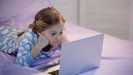 Girl in pajama watching cartoons on laptop in bedroom in evening.