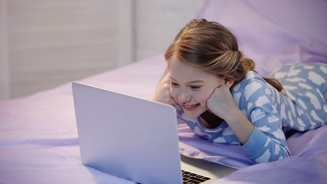 Happy kid in pajama looking at laptop in bedroom in evening.