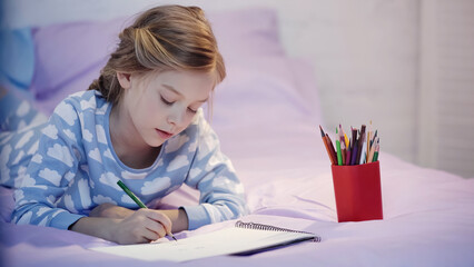 Child in pajama drawing on sketchbook on blurred bed in evening.