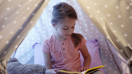 Preteen child in t-shirt reading book in wigwam in evening. © LIGHTFIELD STUDIOS