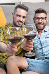 Gay couple in love sitting at home on the floor and toasting with red wine. High quality photography.