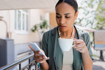 Young african american woman holding cup of cappuccino and using smartphone on terrace of sweet shop.