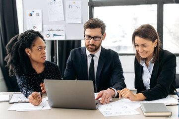 Multiracial colleagues are brainstorming on strategy together, sitting in modern office, studying documents, using laptop, discussing, smiling. Business partners working together on a project