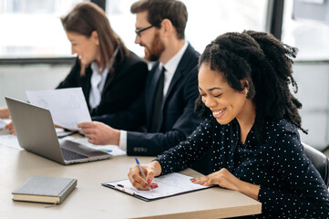 Professional african american young woman in a shirt, a financial manager or ceo, sitting at a desk with her colleagues in the office, concentrated examines the documents, financial report, smiling