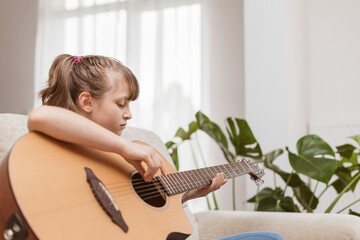 A cute little girl is sitting on the couch at home and learning to play the acoustic guitar