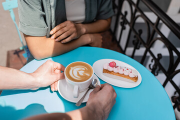 Cropped view of seller putting cappuccino on table near dessert and african american client on terrace of sweet shop.