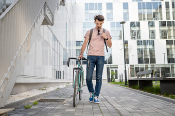 One man young adult male with brown hair and mustaches walking by the building with rucksack on his back and bicycle happy smile joyful real people copy space full length front view