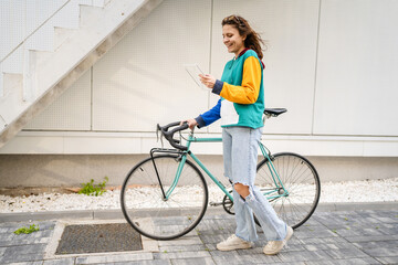 Fototapeta na wymiar One woman young adult caucasian female walking by the university building holding bicycle and digital tablet in day real people copy space happy smile joyful full length side view