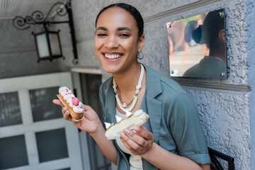 Positive african american customer holding sweet eclairs near confectionery outdoors.