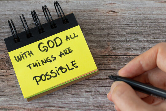 With God all things are possible, handwritten text on a yellow note with a hand on a wooden table. Top view. A closeup.