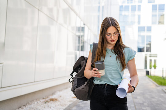 One woman young adult caucasian female student standing and walking in front of university with paper and cup of coffee outdoor in bright day real people copy space
