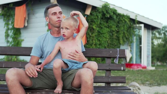 Caucasian father and son are sitting on the beanch in the village. The son is sitting on his father's laps, they are talking and having good time together. Summer in the countryside