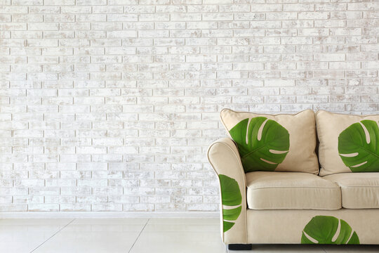 Comfortable sofa with print of tropical monstera leaves near light brick wall in room