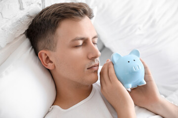 Young man with piggy bank sleeping in bed, closeup