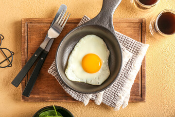 Wooden board with frying pan of fried egg and cutlery on color background