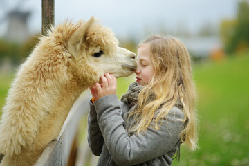 Leuk jong meisje dat een alpaca aait in een boerderijdierentuin op herfstdag. Kind dat een lama voedt op een dierenboerderij. Kid op een kinderboerderij in de herfst.