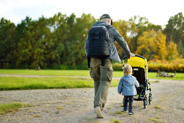 Young father walking in autumn park with toddler son in pushchair. Man pushing a stroller for...