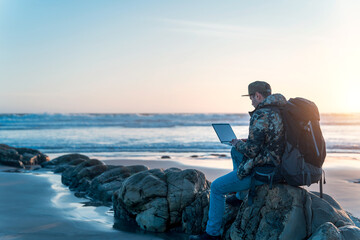 digital nomad sitting on the rock of the  beach working with his laptop at sunset