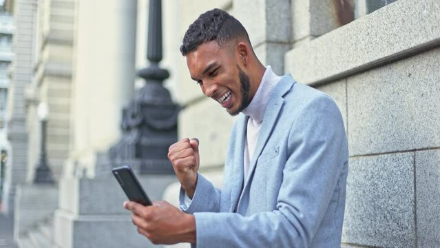 Excited, overjoyed and successful businessman celebrating win with mobile phone outside. Young entrepreneur screaming in happiness at online prize. Fist pumping air with happiness.