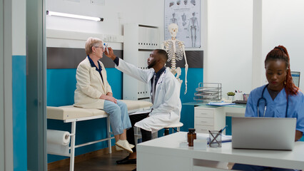 Obraz premium Male physician using thermometer to measure temperature on elderly woman at medical consultation in office. Doctor doing fever measurement with tool on senior patient at cabinet.