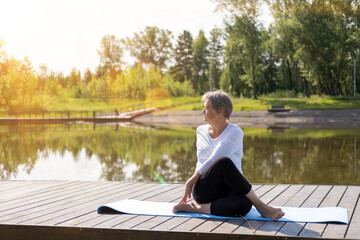 An elderly Caucasian, gray-haired woman does stretching exercises in the park on a yoga mat....