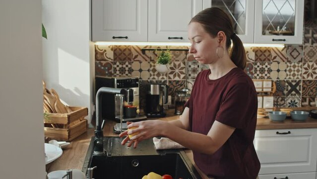 Medium side view of young Caucasian woman rinsing fresh veggies, standing by sink in kitchen in house at daytime