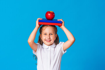 Back to school. cheerful schoolgirl with books on the background of a blue paper wall. little girl is ready to study, holding her school textbooks and red apple on her head. having fun with homework