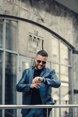 businessman is waiting for a meeting near the office center, looks at the clock.Handsome stylish man in black glasses is smiling.
