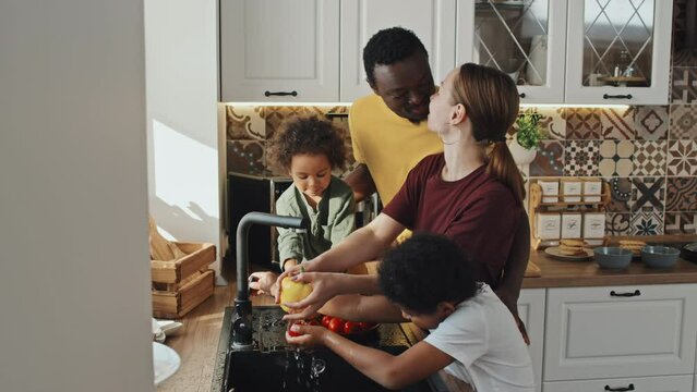 Medium side view of young Caucasian mother of two cute little Black boys rinsing fresh veggies, standing by sink in kitchen in house at daytime, African American man kissing wife