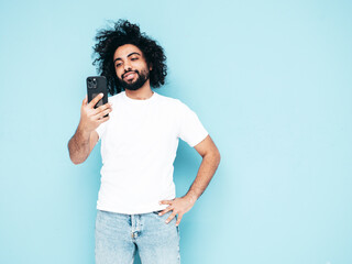 Handsome smiling hipster  model.Sexy Arabian man in summer stylish clothes.Fashion male with long curly hairstyle posing near blue wall in studio.Looking at cellphone screen. Using apps